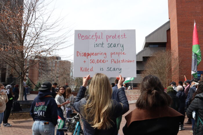 A protester, photographed from behind, carries a sign that reads, "Peaceful protest isn't scary. Disappearing people + 50,000+ Palestinians killed is scary."