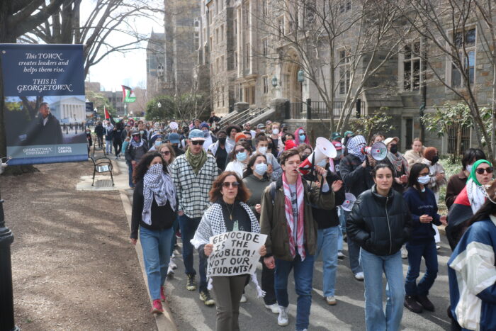 Dozens of protesters walk on the path next to Copley. They walk next to a sign that says "This TOWN creates leaders and helps them rise. This is Georgetown."