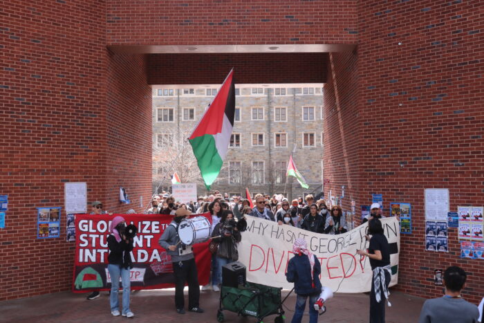 Protesters walk through the archway in Red Square. The protesters in the front are carrying two large banners: one reads, "Globalize the student intifada," while the other reads, "Why hasn't Georgetown divested?" One protesters waves a Palestinian flag on a long flagpole.