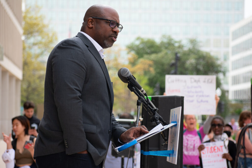 A person wearing a gray suit speaks into a microphone at a podium. Protesters are blurred in the background.