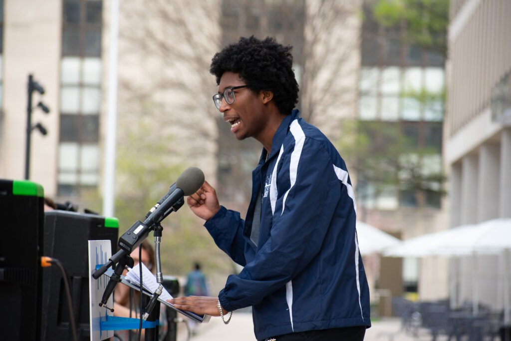 A person wearing a navy jacket speaks at a podium into a microphone, gesturing with a hand.