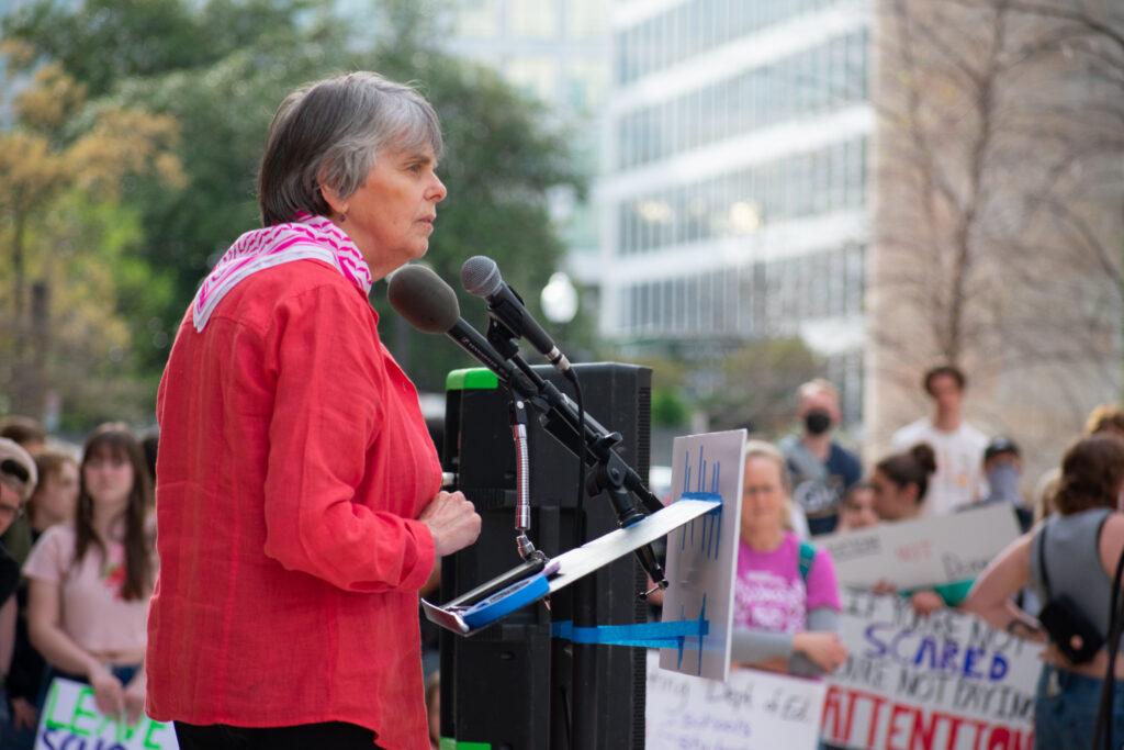 A person in a bright red long-sleeve shirt speaks at a podium to a crowd of people.