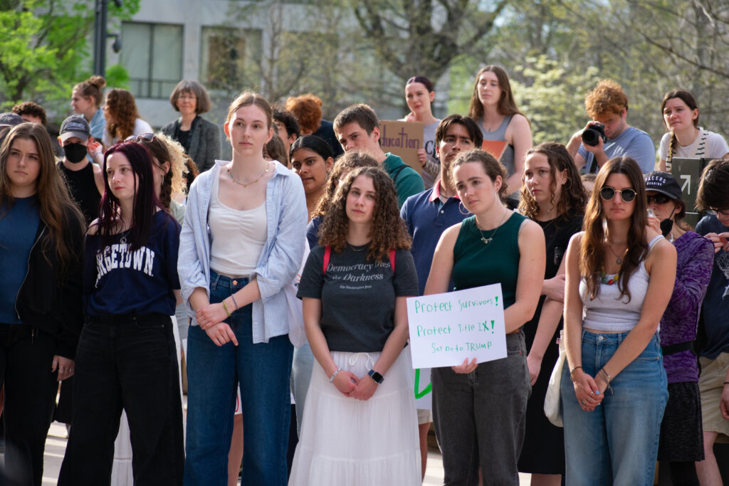 A crowd of people gathers. One person in the front carries a sign reading, "Protect Survivors! Protect Title IX! Say No to Trump"