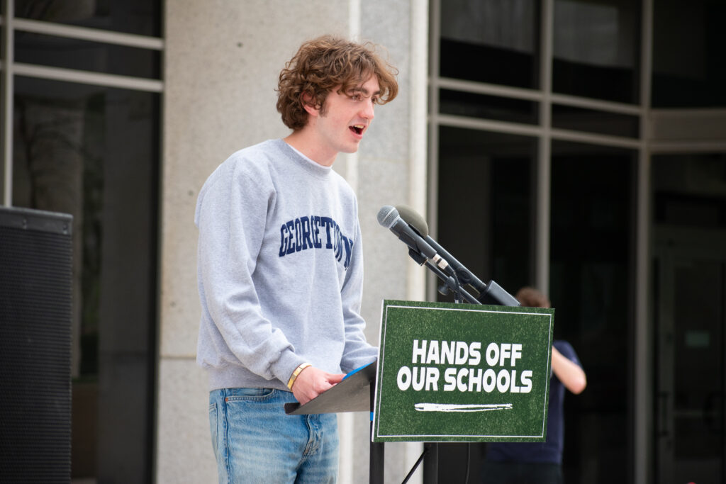 A person speaks at a podium into a microphone. The podium has a green sign on it that reads "HANDS OFF OUR SCHOOLS"