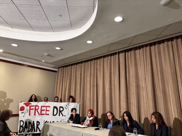 Shows several people lined up at a table against a beige curtain. Next to the table, people hold a sign reading, "FRE DR. BADAR KHAN SURI."