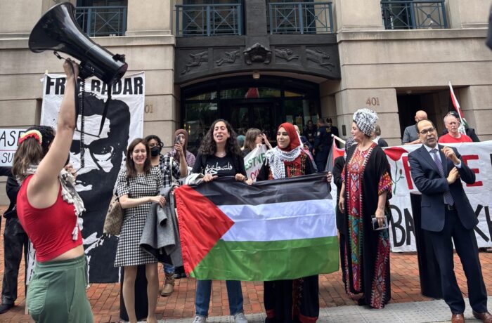 The image shows several people standing outside a courthouse, with one raising a megaphone into the air and two others holding a Palestinian flag