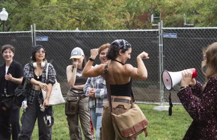 A person wearing a bandana, a black tank top, cargo pants, and a beige sling bag stands in the middle of the picture. They are holding their hands to the side and flexing their muscles. A group of people watch in the background.