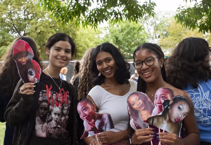 Three smiling young people stand outdoors holding large cutout photos of Mensah-Boone's face in different poses. The person on the left wears a black shirt, the person in the middle wears a white top, and the person on the right wears glasses and holds several cutouts of Mensah-Boone showing different facial expressions. Trees and other people are visible in the background.