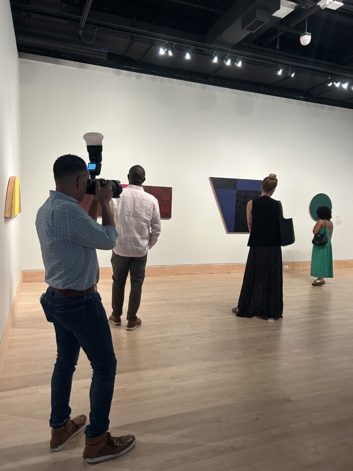 Various people admire art inside the gallery, with one man sporting a large camera with a flash.