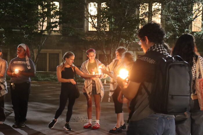 Five people stand in frame. The second person to the left is wearing all black and facing the camera. They hold their hand towards a person on the right, helping them light their candle.