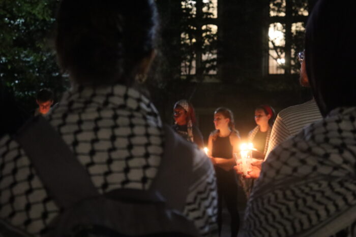 A person wearing a gray backpack and keffiyeh facing away from the camera foregrounds the picture. To their right, there are two people facing the camera looking towards the left of the image. They are both holding a candle.