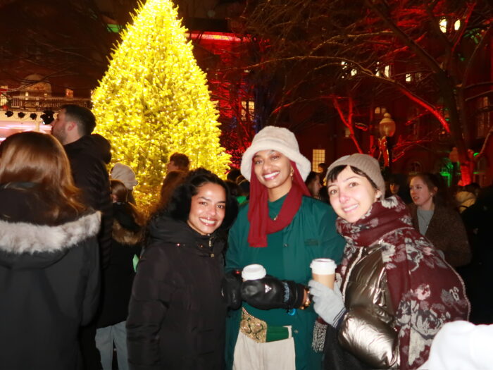 Three women pose in front of the lit Christmas tree.