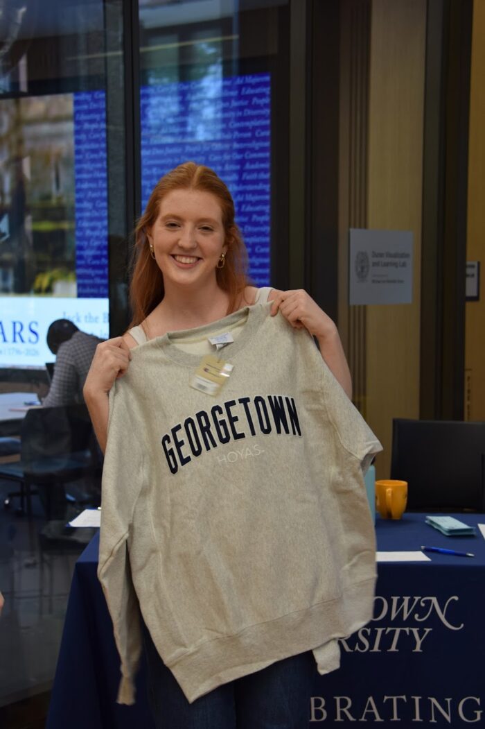 A redheaded student stands smiling with a grey Georgetown crewneck.