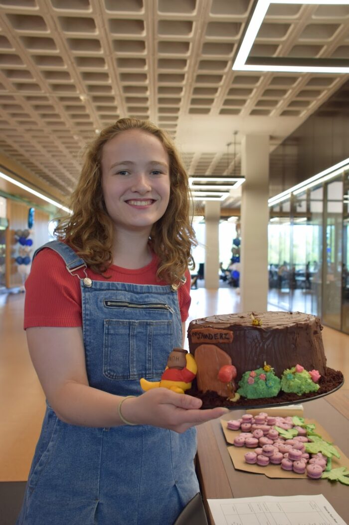 A blonde girl smiles and holds her Winnie the Pooh-inspired brown cake.