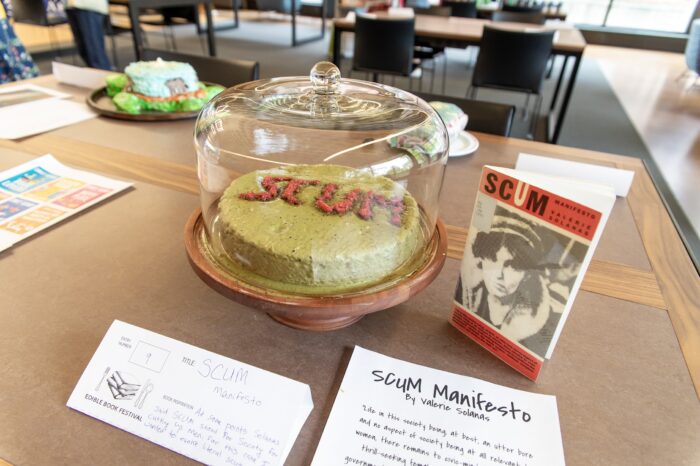 A green cake titled "SCUM" with red frosting, on a brown table.